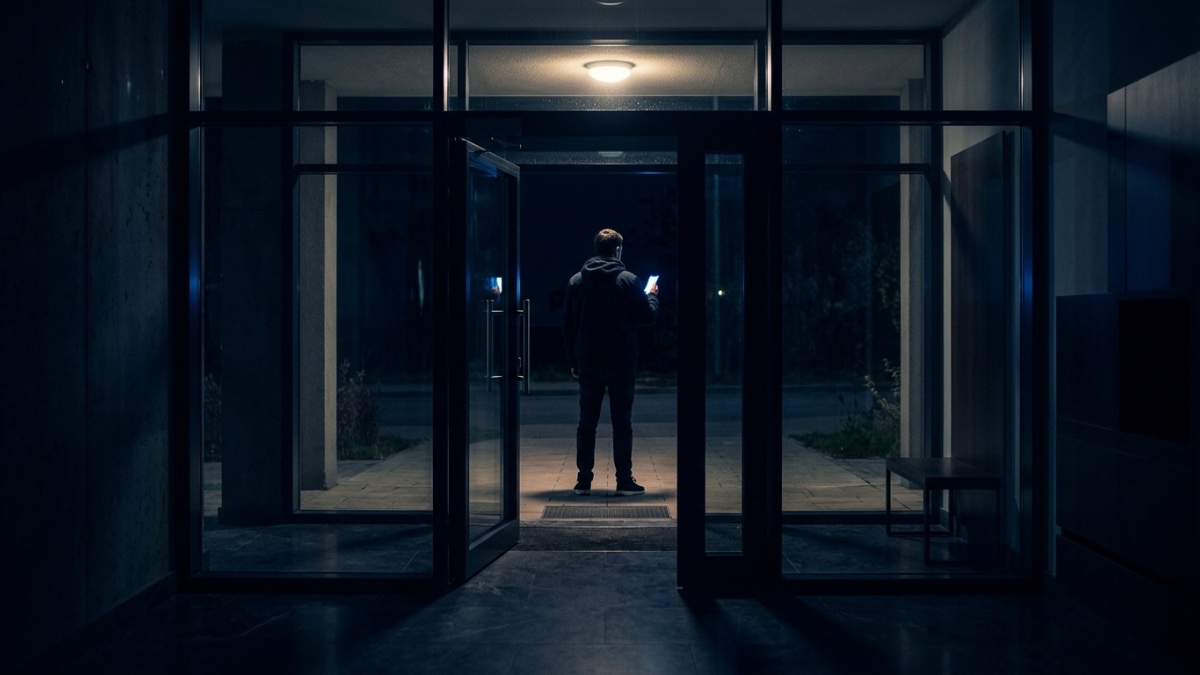 De nuit, vu de l’intérieur du hall d’un immeuble résidentiel, un inconnu attend derrière la porte vitrée, l’écran de son téléphone allumé à la main